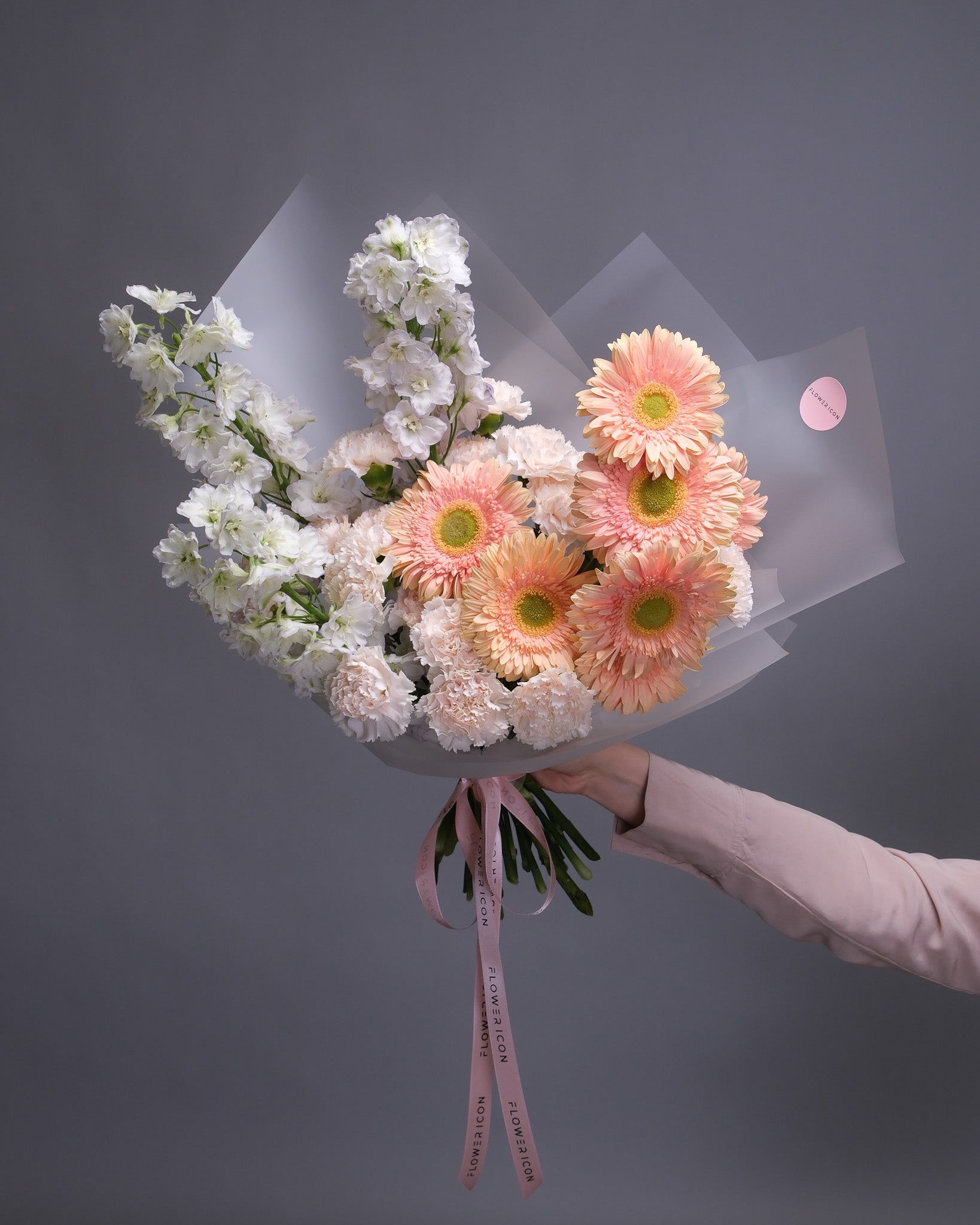 Handheld bouquet in soft blush and peach tones, featuring gerbera daisies, blush carnations, and white delphinium for a light, romantic look.