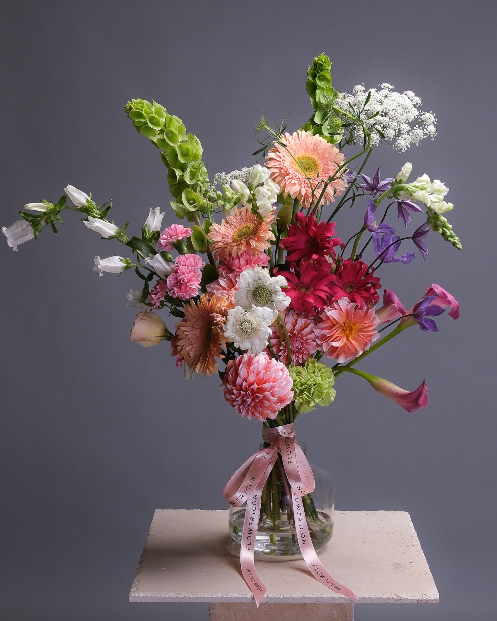 A large bouquet of colorful flowers in a clear vase with a pink ribbon on a gray background