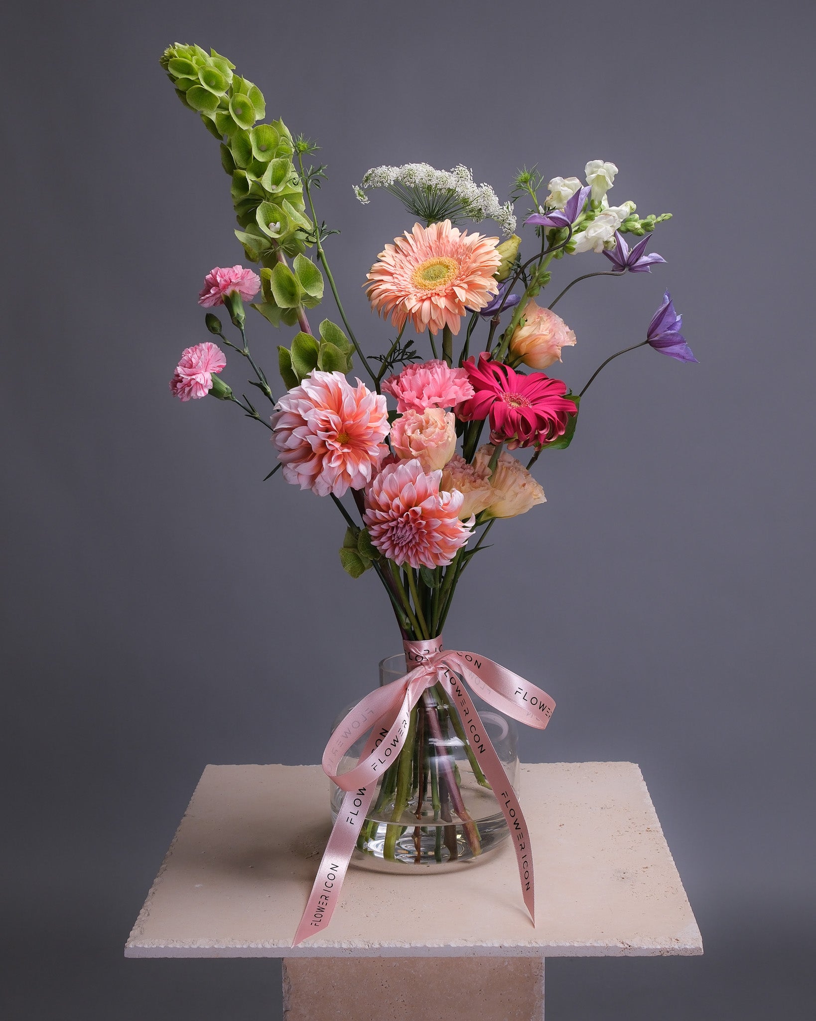 A small Bouquet of colorful flowers in a clear vase with a pink ribbon on a gray background