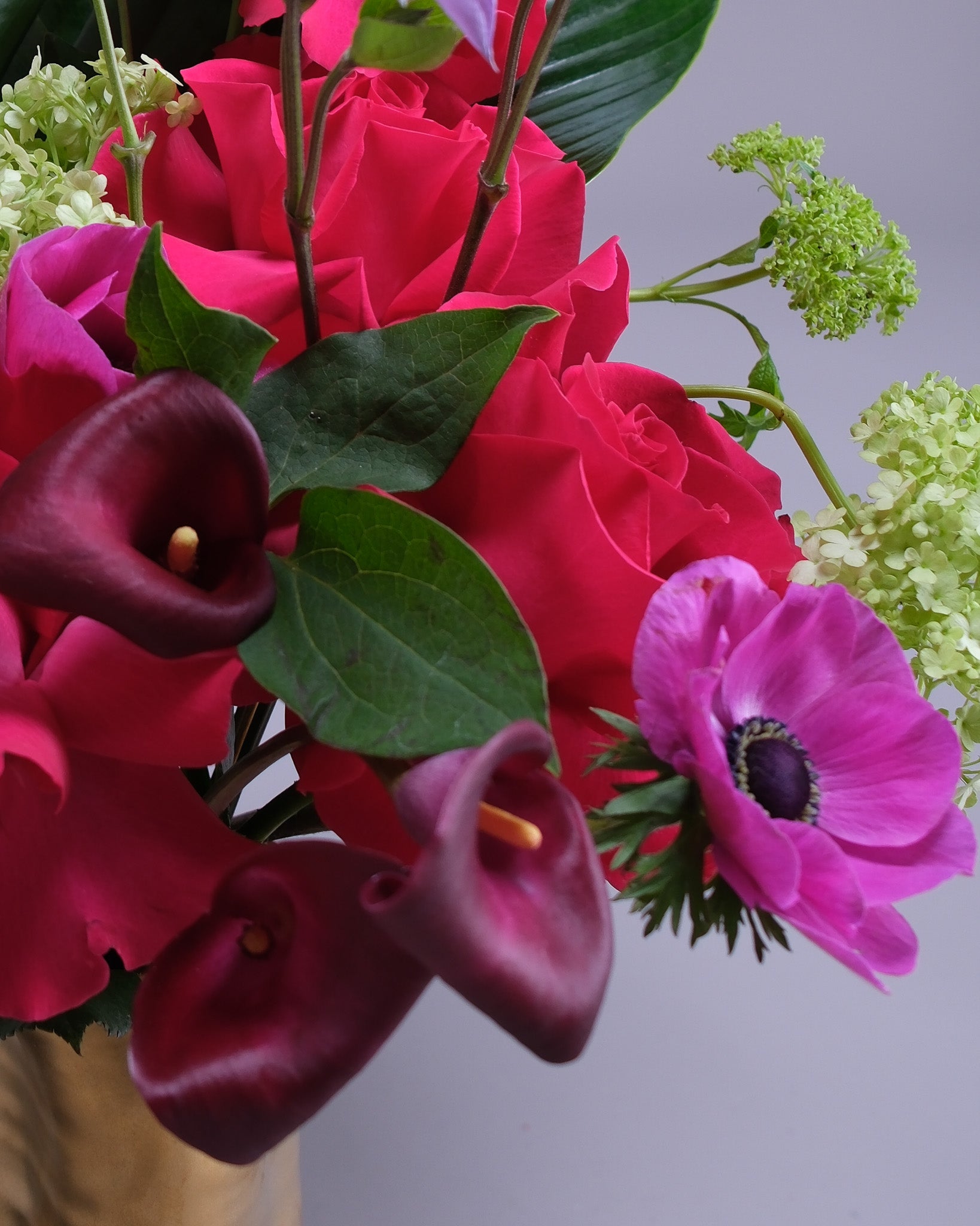 Bouquet of red roses, purple calla lilies, and pink anemones with green leaves on a light gray background.