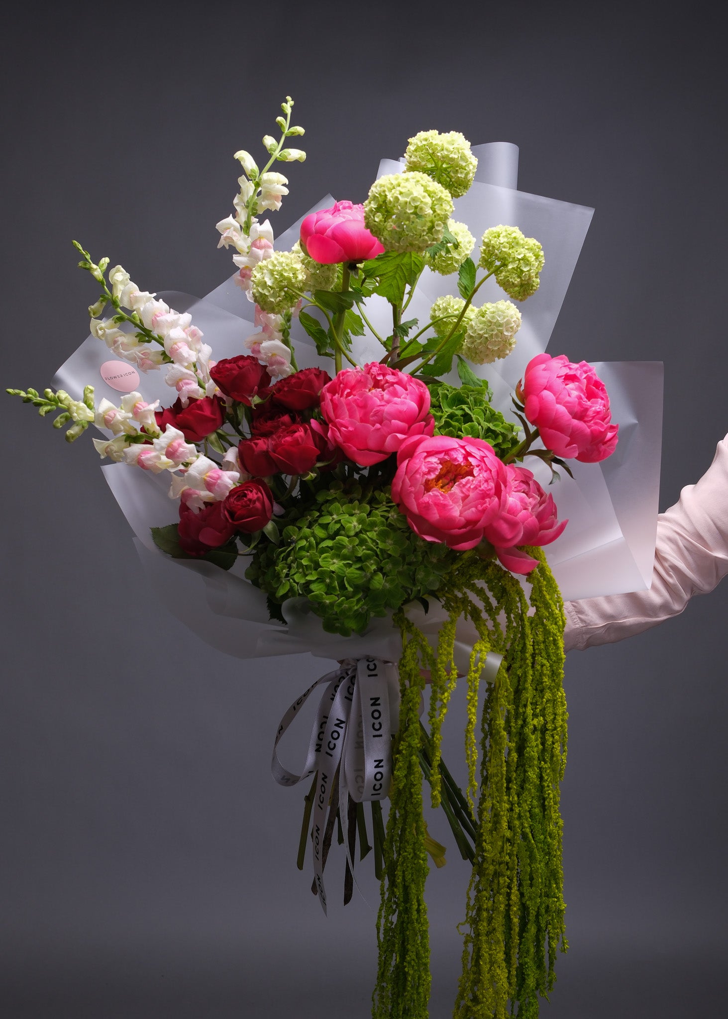 Handheld bouquet featuring green hydrangeas, viburnum, coral peonies, red garden roses, and snapdragons in a lush, oversized arrangement.