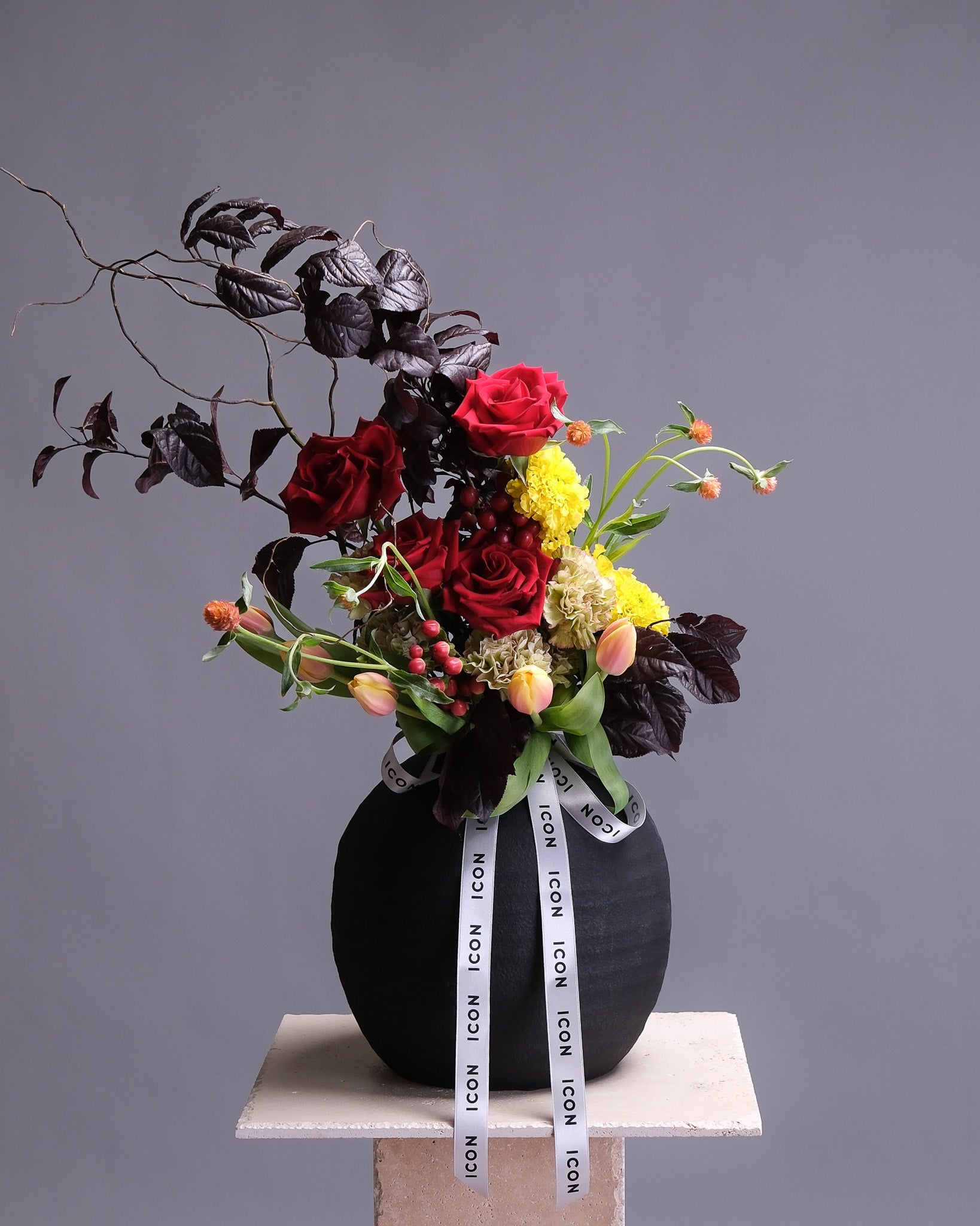 A fall floral arrangement in a black vase with red, yellow, and green flowers on a gray background