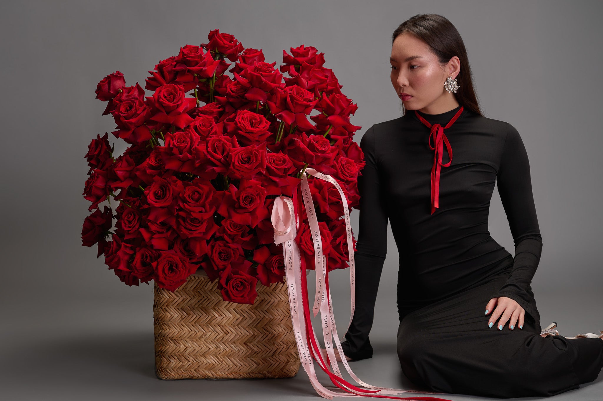 Woman in a black dress with a red ribbon sitting next to a large arrangement of red roses in a woven basket.