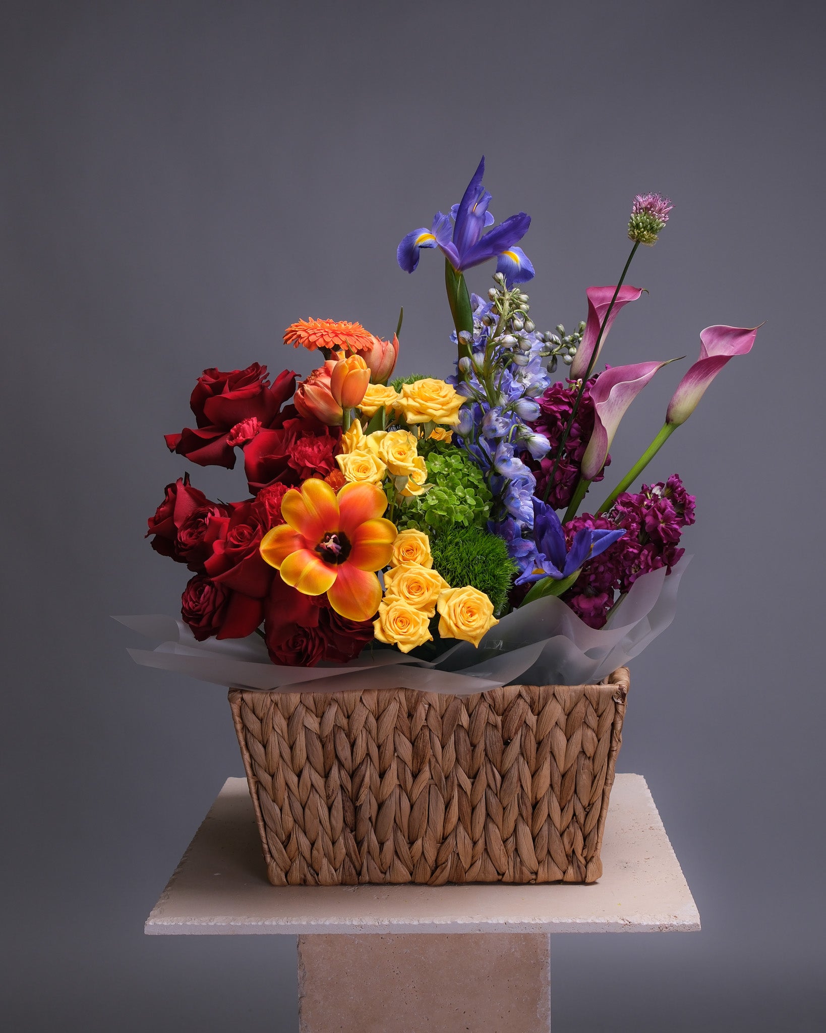 Front view of a rainbow flower arrangement in a woven basket, featuring vibrant blooms in every color including roses, tulips, hydrangeas, and orchids.