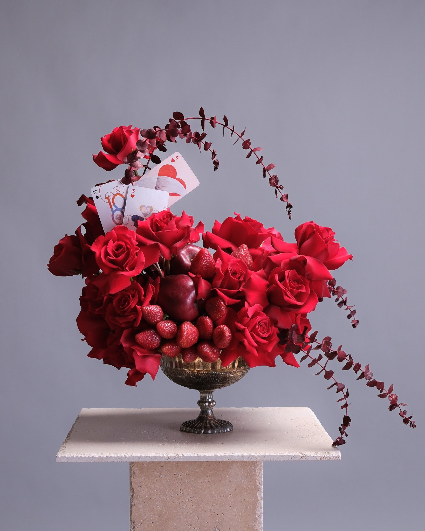 Decorative arrangement of red roses, strawberries, and playing cards in a silver vase on a gray background.